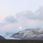 A bright blue glacial lagoon surrounded by gray mountains, with a full moon rising in the background